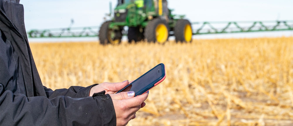 Woman's hands holding a smartphone with a tractor with sprayer attachment in a harvested field in the far background